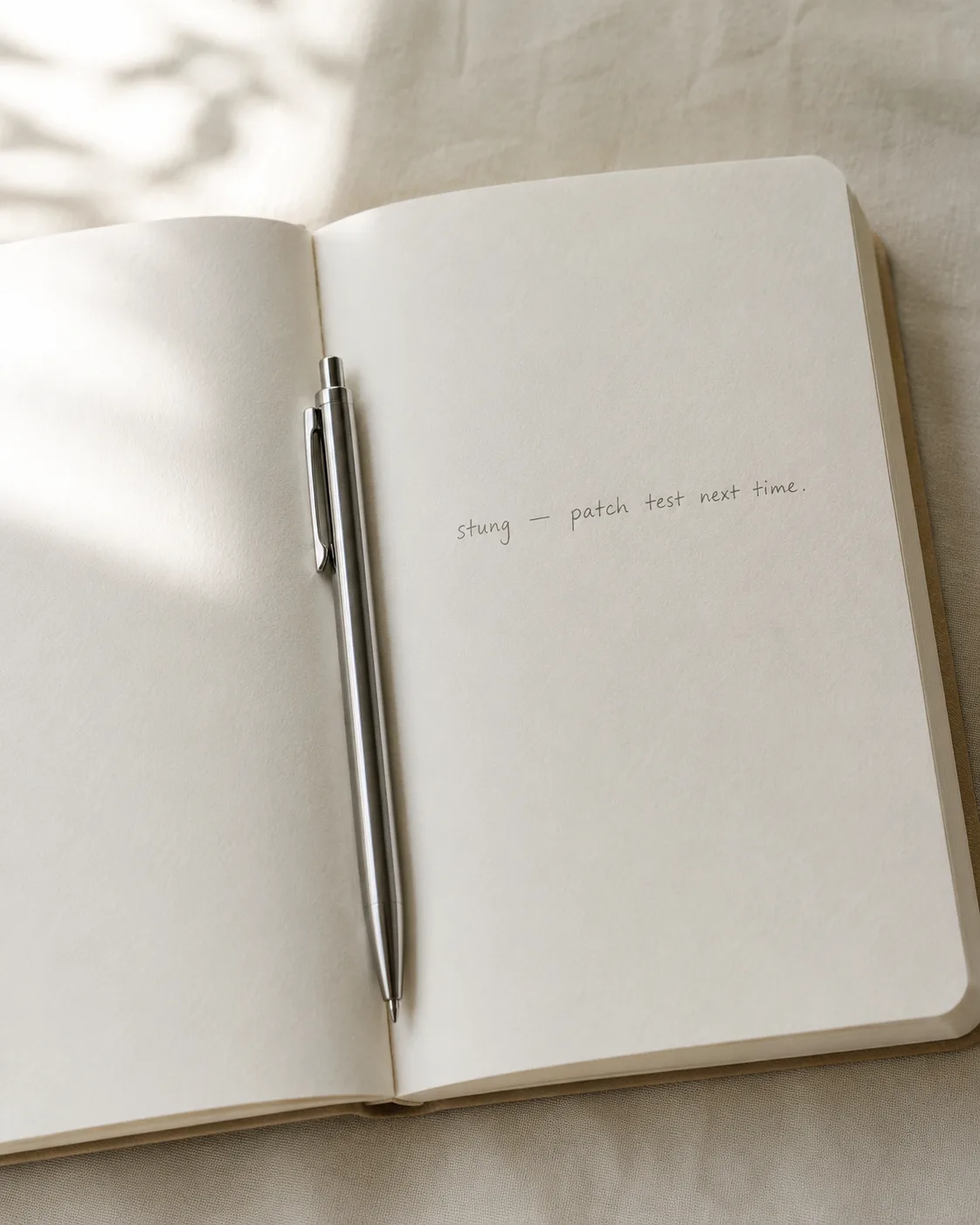 An open notebook on linen with a slim ballpoint pen, faint handwritten note reading 'stung — patch test next time' in soft daylight.